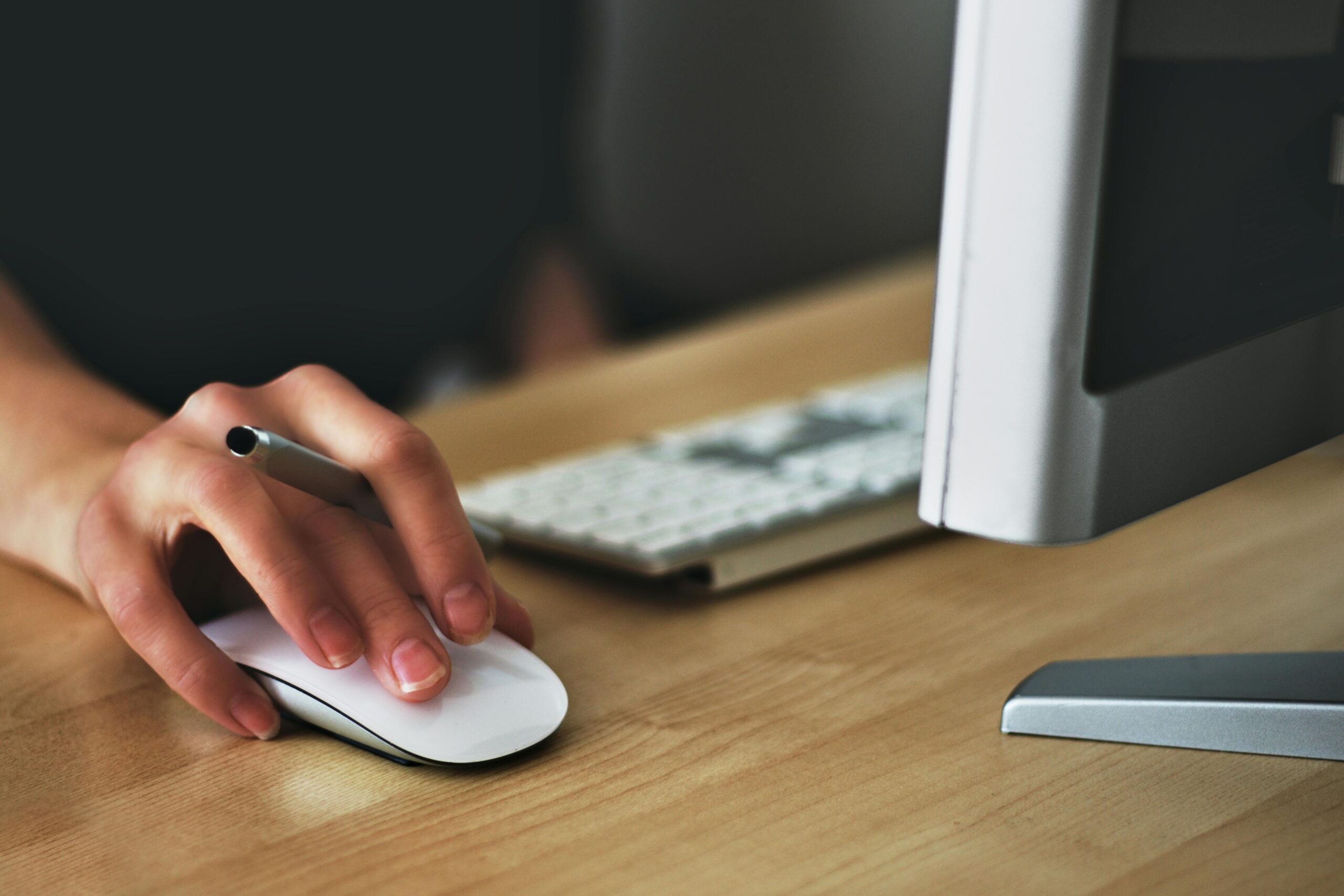 A hand using a wireless mouse at a modern desk setup with a computer and keyboard.